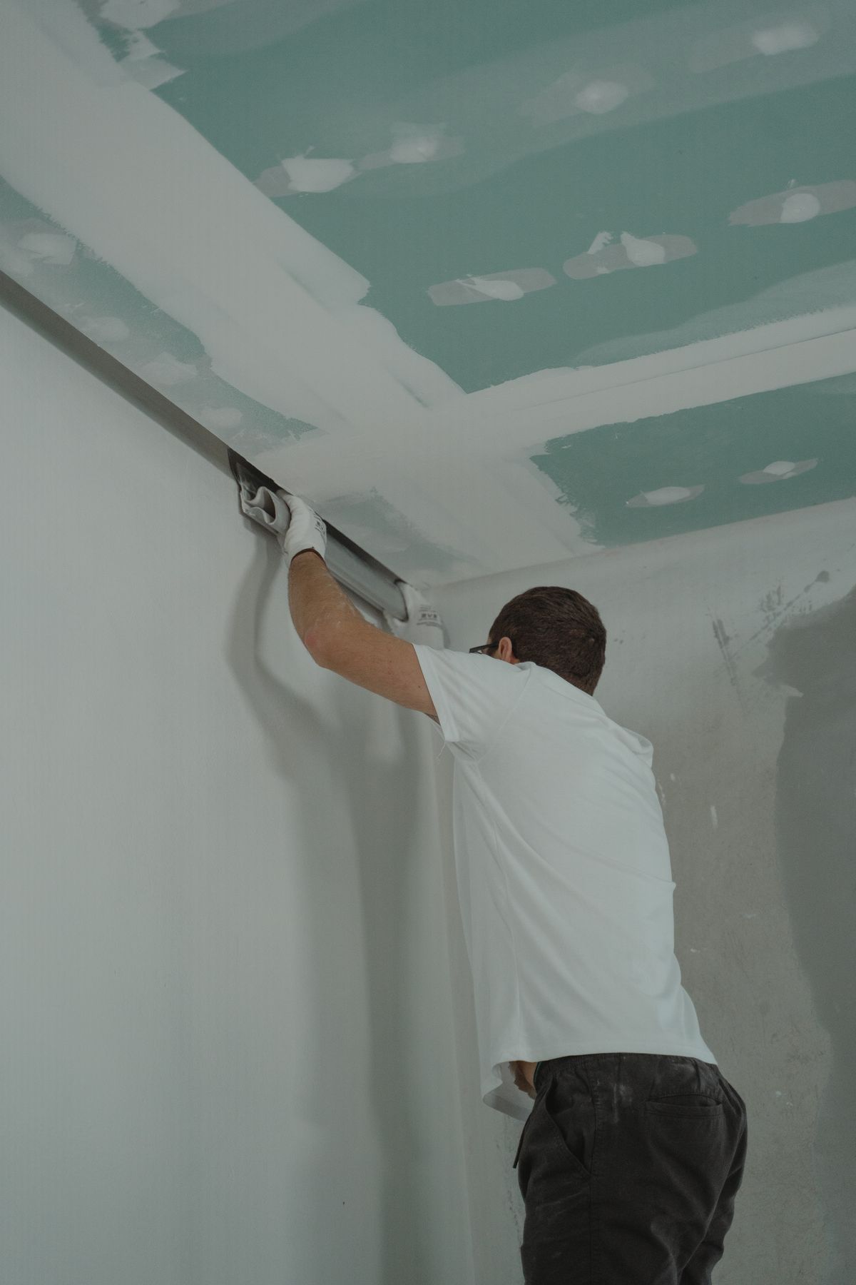 Worker pulling a wide finishing knife along a ceiling-to-wall corner joint on fresh drywall